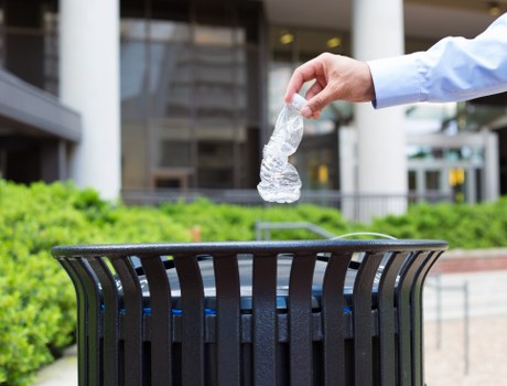 A close-up of a person's hand wearing a light blue long-sleeved shirt as they dispose of a crumpled clear plastic water bottle into a black outdoor waste bin with vertical slats. The waste bin is situated on a paved area with a background featuring a modern building with glass windows and concrete columns, indicating an urban or commercial environment possibly in Elephant and Castle. The scene is well-lit, suggesting daytime, and there is some greenery visible near the base of the building, contributing to a tidy outdoor setting. The image emphasizes proper rubbish disposal as part of waste management services offered by Commercial Waste Elephant and Castle in the local area.