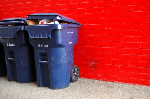 Two dark blue plastic wheelie bins with black lids are positioned on a paved surface beside a red brick wall. The bins are filled with mixed waste, visible in the open-top sections, including cardboard boxes and plastic packaging. The bins are placed close to each other, with one slightly in front of the other, and both have textured surfaces with ridges and identification labels on the sides. The wheels are underneath, and the background features a vibrant red brick wall with evenly spaced bricks and mortar lines, suggesting a commercial or residential area in Elephant and Castle. The lighting is natural, casting minimal shadows, and the scene reflects typical rubbish collection or removal preparation, aligning with local waste management services provided by Commercial Waste Elephant and Castle.