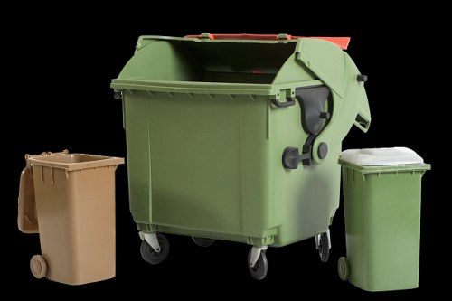The image shows three different wheeled rubbish bins against a black background. The largest bin in the centre is a green plastic wheelie bin with a closed lid, featuring a textured surface, side handles, and locking mechanisms on the side. To the left, there is a smaller beige bin with a smooth surface, a lid, and two small wheels at the base, positioned slightly behind the larger green bin. On the right, a slightly taller green bin with a flat lid is visible, also with a smooth texture and small wheels. The scene appears to depict waste collection containers, suitable for commercial or residential rubbish disposal, with a neutral lighting setup highlighting textures and colours.