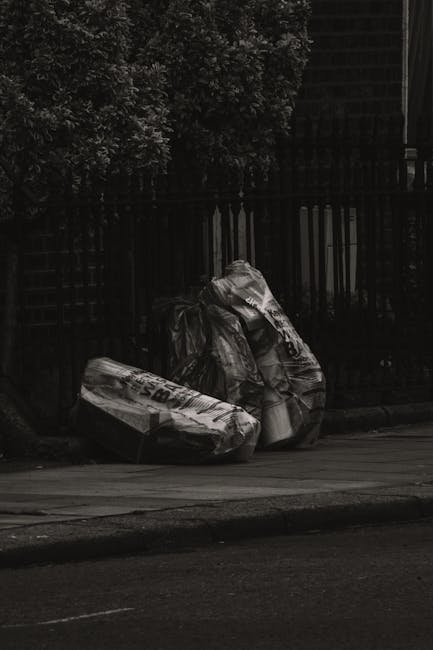 A group of large black garbage bags, made of crinkled plastic material, are positioned on the pavement adjacent to a black metal fence and a sidewalk. The bags appear to contain waste or discarded ite