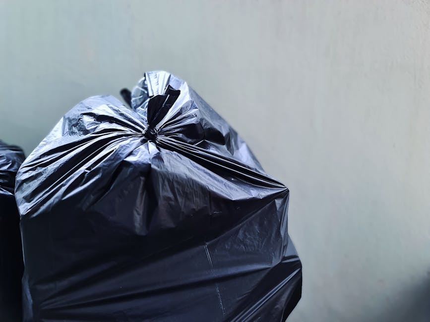 A group of large black garbage bags, made of crinkled plastic material, are positioned on the pavement adjacent to a black metal fence and a sidewalk. The bags appear to contain waste or discarded ite